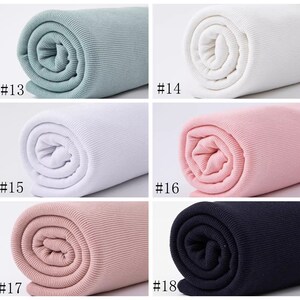 Op de afbeelding: Close-up van zes rollen geribbelde stof in verschillende kleuren: lichtblauw, wit, roze, lichtroze en zwart. De stof is opgerold en de uiteinden zijn zichtbaar. De nummers #13, #14, #15, #16, #17 en #18 zijn onder elke rol afgedrukt.