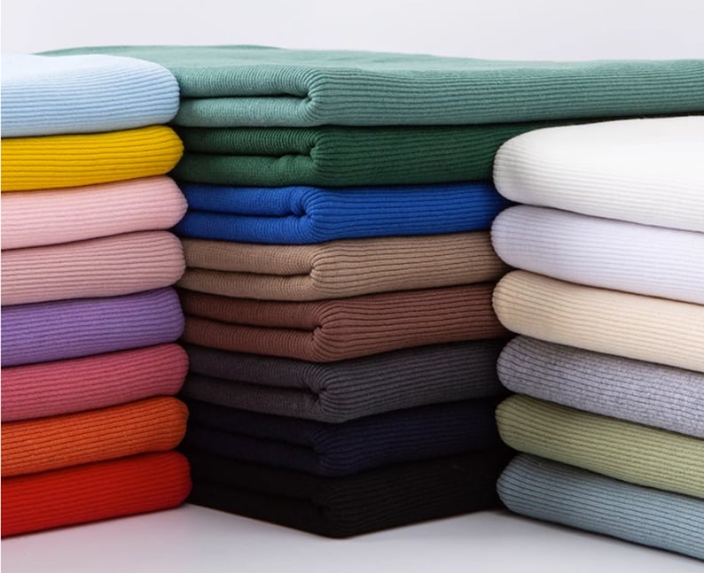 Op de afbeelding: Een verscheidenheid aan geribbelde breisels in verschillende kleuren, waaronder lichtblauw, geel, roze, paars, rood, oranje, groen, wit, beige, grijs en blauw. De breisels zijn gestapeld in stapels, waarbij elke stapel een andere kleur bevat.
