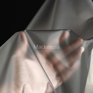 May include: A sheet of translucent grey fabric is held up, with a hand visible beneath. The fabric has a subtle sheen and is draped in soft folds. The word "Mackenziu" is printed on the fabric.