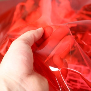 May include: A close-up of a hand holding a bright red, transparent plastic sheet. The sheet is folded over, revealing the texture of the plastic.