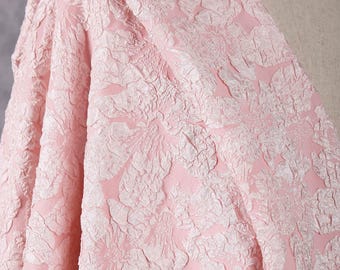 May include: A close-up of a pink fabric with a raised floral pattern. The fabric is draped over a form, showcasing its texture and design. The color is a soft, light pink with a subtle sheen, suitable for apparel or home decor projects.