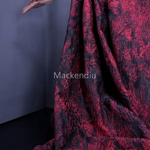 Puede incluir: Una tela drapeada con un patrón texturizado en tonos rojos y negros. La tela está drapeada sobre un soporte de madera. El texto "Mackendiu" es visible en la tela.