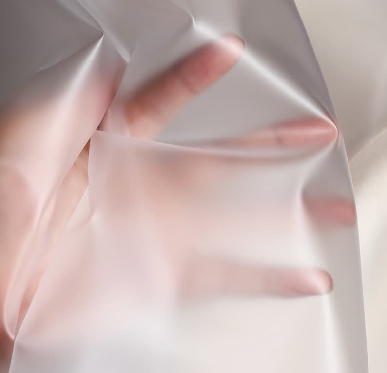 May include: A translucent, white fabric with a smooth, silky texture. The fabric is draped over a hand, creating soft folds and wrinkles.