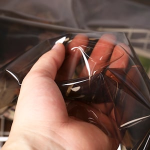 May include: A hand holding a sheet of dark, transparent plastic. The plastic has a reflective quality, with highlights and shadows creating a sense of depth. The material appears to be thin and flexible.