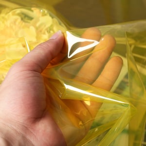 Puede incluir: Primer plano de una mano sosteniendo una lámina de plástico transparente amarillo. El plástico es translúcido y tiene un ligero brillo.