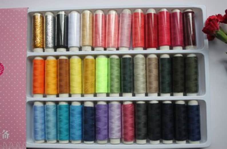 K&ouml;nnte beinhalten: Ein Set mit 36 Garnrollen in verschiedenen Farben, darunter Gold, Silber, Rot, Gr&uuml;n, Blau und Lila. Die Rollen sind in einem Plastikkoffer mit einem durchsichtigen Deckel angeordnet.