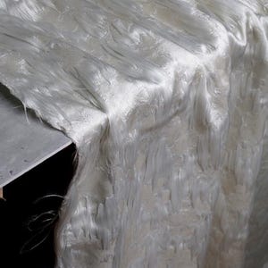 Könnte beinhalten: Nahaufnahme eines weißen, strukturierten Stoffes mit geschichtetem, faserigem Aussehen. Das Material scheint über einer Oberfläche drapiert zu sein, mit sichtbaren Falten und einer weichen, schimmernden Qualität. Die Textur des Stoffes deutet auf eine mögliche Verwendung in Kleidung oder Heimdekoration hin.