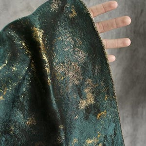 Puede incluir: Primer plano de una tela verde oscuro con una superficie texturizada. La tela está adornada con detalles metálicos dorados, creando un efecto brillante. El material se sostiene, revelando su caída y textura.