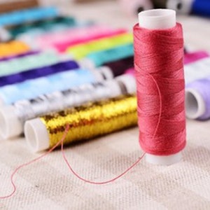 K&ouml;nnte beinhalten: Nahaufnahme einer Spule mit pinkfarbenem Garn, im Hintergrund sind verschiedene andere Garnrollen zu sehen. Die Spulen sind in einer Reihe angeordnet und haben verschiedene Farben, darunter Gold, Blau, Lila und Gr&uuml;n.
