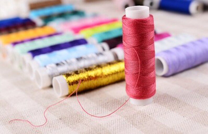 K&ouml;nnte beinhalten: Nahaufnahme einer Spule mit pinkfarbenem Garn, im Hintergrund sind verschiedene andere Garnrollen zu sehen. Die Spulen sind in einer Reihe angeordnet und haben verschiedene Farben, darunter Gold, Blau, Lila und Gr&uuml;n.