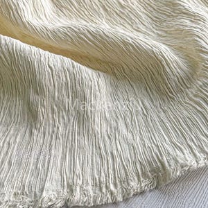 Könnte beinhalten: Nahaufnahme eines cremefarbenen Stoffes mit einer strukturierten, zerknitterten Oberfläche. Der Stoff hat einen rohen, fransigen Rand und eine subtile, natürliche Farbvariation. Das Material scheint leicht und fließend zu sein.