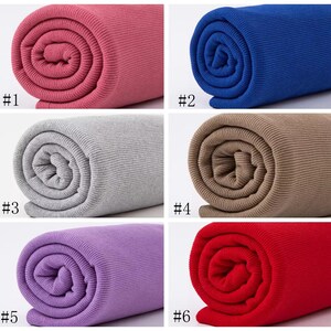 Op de afbeelding: Zes rollen geribbelde stof in verschillende kleuren: roze, blauw, grijs, bruin, paars en rood. De stof is opgerold en de uiteinden zijn zichtbaar.