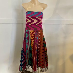 May include: A vibrant strapless dress displayed on a mannequin. The bodice has a geometric design in pink, teal, and beige. The skirt is a patchwork of patterned fabrics in red, orange, and teal, finished with a fringed hem.