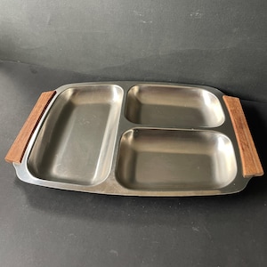 May include: A rectangular stainless steel serving tray featuring three distinct compartments for food organization. The tray includes two smooth, light brown wooden handles attached to the left and right sides, providing a contrast to the metallic finish.