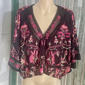 May include: Black V-neck top with pink floral embroidery. The top features a sheer neckline and sleeves, with a cropped design. The floral pattern includes pink flowers and green leaves, creating a bohemian style.