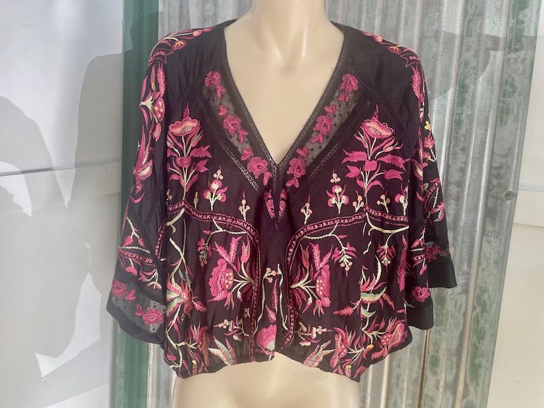 May include: Black V-neck top with pink floral embroidery. The top features a sheer neckline and sleeves, with a cropped design. The floral pattern includes pink flowers and green leaves, creating a bohemian style.
