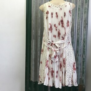 Puede incluir: Un vestido blanco sin mangas con estampado floral y una superposici&oacute;n de encaje. El vestido tiene una falda plisada y un lazo en la cintura.