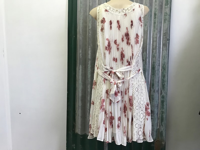 Puede incluir: Un vestido blanco sin mangas con estampado floral y una superposici&oacute;n de encaje. El vestido tiene una falda plisada y un lazo en la cintura.