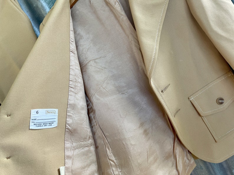 May include: A beige blazer with a pocket on the right side. The inside of the blazer is a light beige color. The label on the inside of the blazer says "Sears" and "6". The label also says "Machine Wash Warm, Tumble Dry Low."