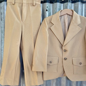 May include: A tan suit with a blazer and pants. The blazer has two flap pockets and three buttons. The pants have a wide leg.
