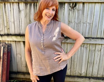 May include: A woman wearing a sleeveless, light brown linen top with a button-up front and black trousers. She is standing in front of a wooden fence.