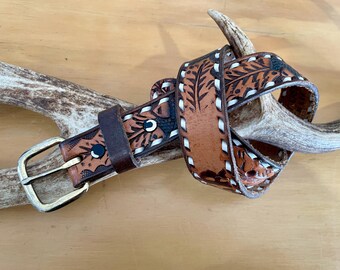 May include: A brown leather belt with a decorative floral pattern and white stitching. The belt has a silver-coloured buckle and is wrapped around a deer antler. The belt is approximately 3.8 cm wide.