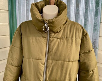 Könnte beinhalten: Eine grüne Steppjacke mit hohem Kragen und Reißverschluss. Die Jacke hat einen silbernen Ringzug am Reißverschluss.