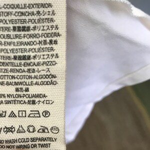Puede incluir: Una etiqueta de tela blanca con instrucciones de cuidado para una prenda de vestir. La etiqueta incluye texto en varios idiomas, incluidos ingl&eacute;s, franc&eacute;s, alem&aacute;n, italiano, espa&ntilde;ol y japon&eacute;s. La etiqueta tambi&eacute;n incluye una serie de s&iacute;mbolos que indican c&oacute;mo cuidar la prenda, como lavar a mano con agua fr&iacute;a, no retorcer ni escurrir, y no blanquear.