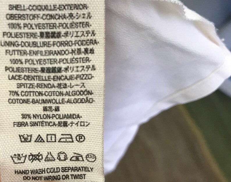 Puede incluir: Una etiqueta de tela blanca con instrucciones de cuidado para una prenda de vestir. La etiqueta incluye texto en varios idiomas, incluidos ingl&eacute;s, franc&eacute;s, alem&aacute;n, italiano, espa&ntilde;ol y japon&eacute;s. La etiqueta tambi&eacute;n incluye una serie de s&iacute;mbolos que indican c&oacute;mo cuidar la prenda, como lavar a mano con agua fr&iacute;a, no retorcer ni escurrir, y no blanquear.