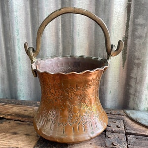 Könnte beinhalten: Ein Kupfer-Eimer mit einem Messinggriff. Der Eimer hat einen gewellten Rand und ist mit einem geprägten Blumenmuster verziert.