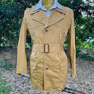 May include: A tan coloured jacket with a belt and two pockets. The jacket has a button closure and a collar.
