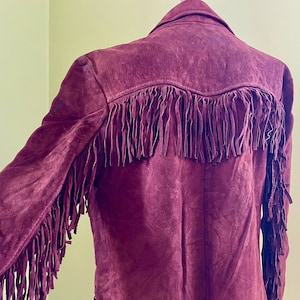 May include: A burgundy suede jacket with fringe detailing on the sleeves and back. The jacket has a classic Western style.