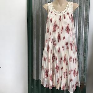Puede incluir: Un vestido blanco sin mangas con estampado floral y escote de encaje. El vestido es plisado y tiene una silueta fluida.