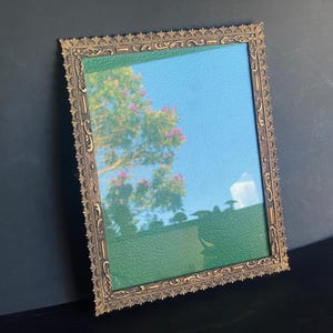 Puede incluir: Espejo antiguo rectangular con un marco dorado ornamentado. El espejo refleja un cielo azul, un árbol con flores rosas y una superficie verde texturizada. El marco tiene un patrón decorativo detallado.