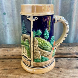 Può includere: Una boccale di birra in ceramica con un design blu e bianco che presenta una scena di paesaggio con alberi e un castello. La boccale ha un manico e le parole "Rotsparti" e "Ruine" sono visibili sul fondo.