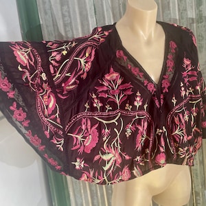 May include: Black embroidered top with a V-neck and wide sleeves. The top features a floral design in shades of pink and cream. The fabric appears to be lightweight, with a sheer lace trim along the edges. The top is displayed on a mannequin.