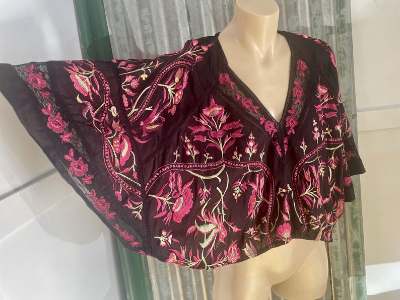 May include: Black embroidered top with a V-neck and wide sleeves. The top features a floral design in shades of pink and cream. The fabric appears to be lightweight, with a sheer lace trim along the edges. The top is displayed on a mannequin.