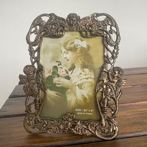 Puede incluir: Un marco de fotos de metal dorado adornado con un diseño floral y acentos de querubines. El marco contiene una fotografía vintage de una mujer sosteniendo flores. El marco mide 3 1/2" x 5" y está hecho en Taiwán.