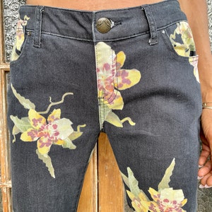 May include: Black denim jeans with a floral print featuring yellow, white, and purple orchids. The jeans have a classic five-pocket style and a button closure.