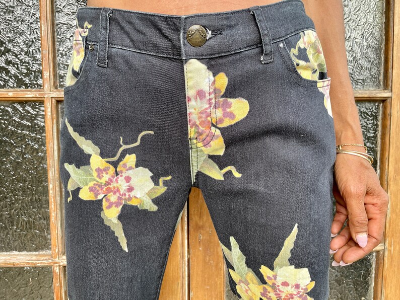 May include: Black denim jeans with a floral print featuring yellow, white, and purple orchids. The jeans have a classic five-pocket style and a button closure.