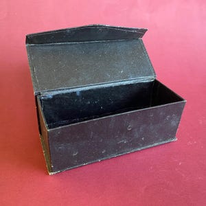 May include: A black rectangular box with an open lid. The box is made of a dark material, possibly cardboard, with a slightly worn texture. The box is positioned on a red surface.