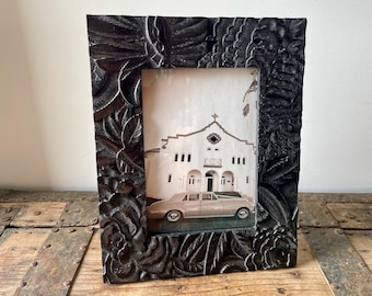May include: A black wooden picture frame with an ornate floral design. The frame is holding a photograph of a white building with a car parked in front of it.