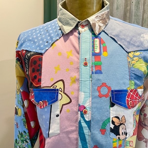 May include: A patchwork button-down shirt with a variety of cartoon characters and patterns. The shirt features a light blue polka dot shoulder, a pink center with yellow stars, and a blue pocket with a cartoon character. The collar is a light gray and brown.