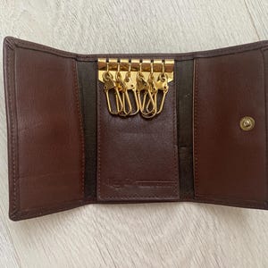 May include: A brown leather key holder with a fold-over flap and a snap closure. The key holder has six gold key rings.