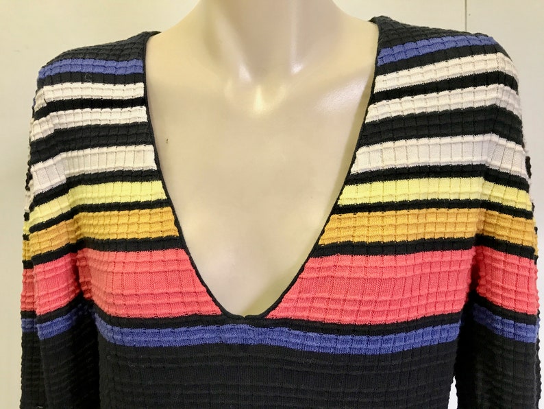 Puede incluir: Un su&eacute;ter de rayas blancas y negras con rayas horizontales de colores en tonos azules, amarillos, naranjas y rosas. El su&eacute;ter tiene un cuello en V.