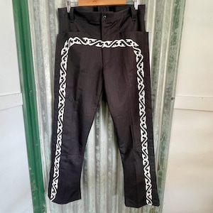 May include: Black trousers with white decorative trim. The trousers are hanging on a wooden hanger. The white trim is a repeating pattern that runs along the sides and across the top of the trousers.