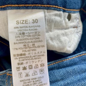 May include: A white tag with black text detailing the size, fabric composition, and care instructions for a pair of blue jeans. The tag reads "SIZE: 30", "49% RAYON-RAYONNE-VISCOSA-RAIOM-", "32% COTTON-COTON-ALGOD&Oacute;N-COTONE-BAUMWOLLE-ALGOD&Atilde;O-", "17% POLYESTER-POLI&Eacute;STER-POLIESTERE", "2% SPANDEX-ELASTHANNE-ELASTANO-ELASTAN-", "MACHINE WASH COLD", "DO NOT BLEACH", "TUMBLE DRY LOW", "IF NEEDED".