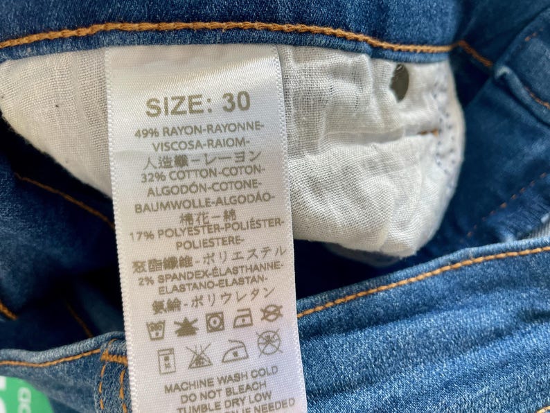 May include: A white tag with black text detailing the size, fabric composition, and care instructions for a pair of blue jeans. The tag reads "SIZE: 30", "49% RAYON-RAYONNE-VISCOSA-RAIOM-", "32% COTTON-COTON-ALGOD&Oacute;N-COTONE-BAUMWOLLE-ALGOD&Atilde;O-", "17% POLYESTER-POLI&Eacute;STER-POLIESTERE", "2% SPANDEX-ELASTHANNE-ELASTANO-ELASTAN-", "MACHINE WASH COLD", "DO NOT BLEACH", "TUMBLE DRY LOW", "IF NEEDED".