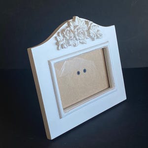 May include: A white rectangular picture frame with a decorative floral design at the top. The frame has a curved top and a rectangular opening for a photo. The frame is made of a solid material.