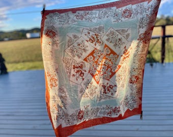 Könnte beinhalten: Ein quadratischer Schal mit floralem Muster in Rot-, Orange- und Cremetönen auf hellblauem Hintergrund. Der Schal mit rotem Rand hängt draußen und weist ein sich wiederholendes Blumenmuster auf.